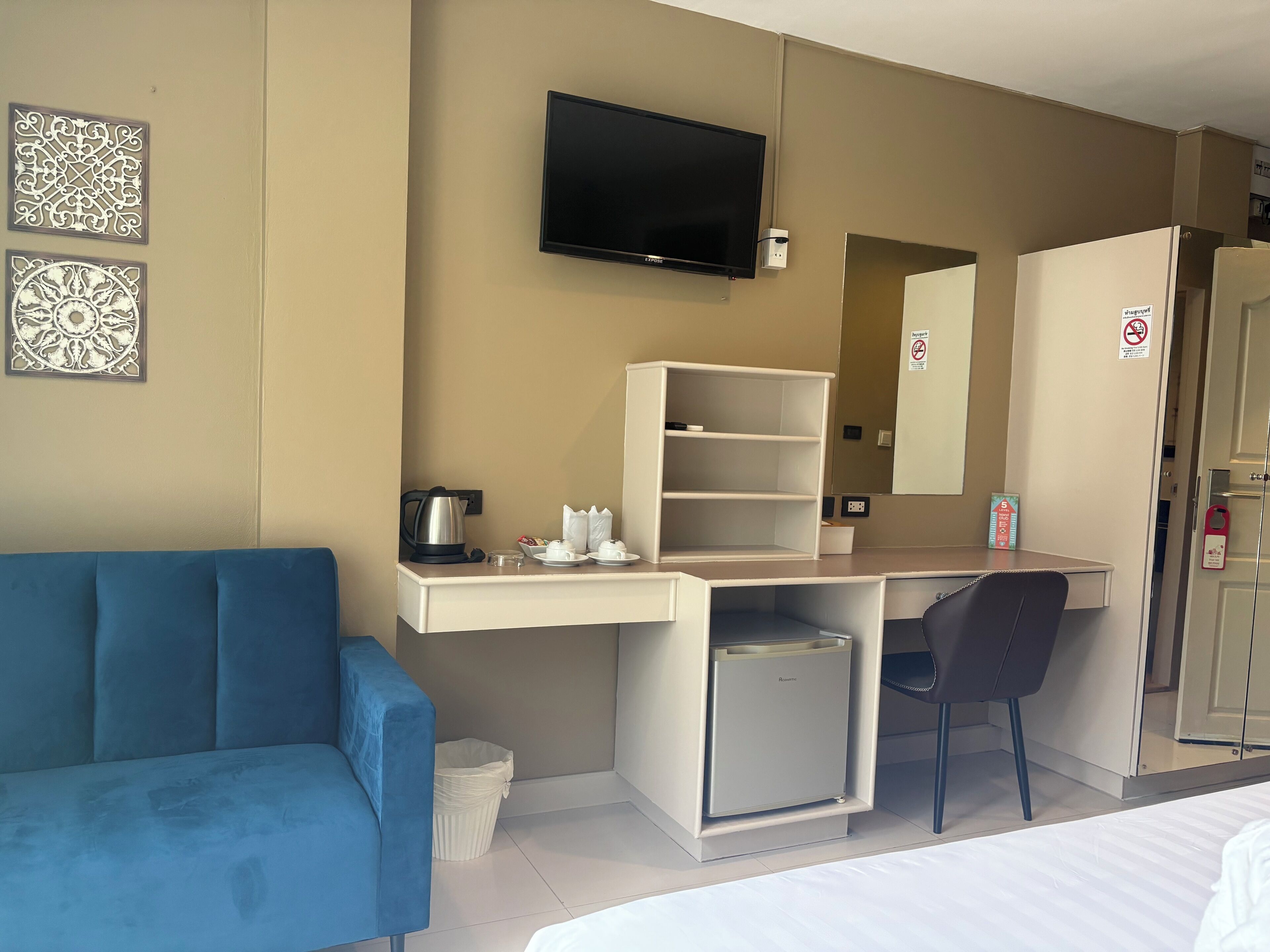 A hotel room with a blue sofa, a desk with a television, a small refrigerator, and a trash bin.