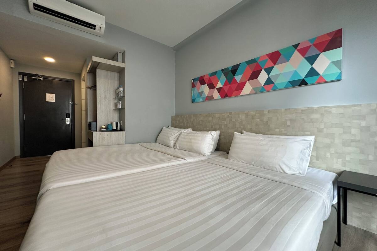 Una camera d'albergo con un letto grande, un condizionatore d'aria a parete, un'opera d'arte geometrica colorata e un pavimento in legno.