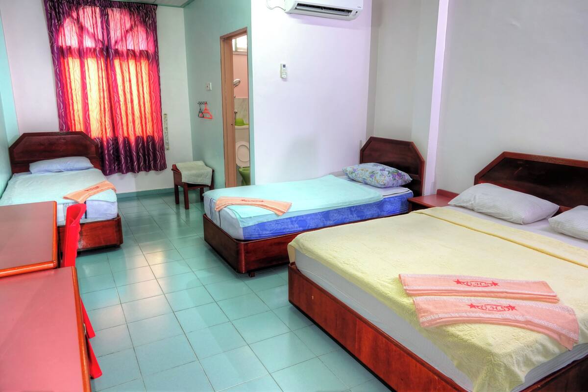 A hotel room with three beds, a desk, and a chair. There is a bathroom visible through an open door.