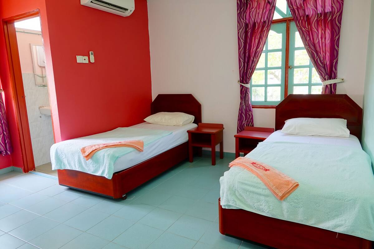 A room with two wooden beds, each with a white pillow and a light blue bedspread. There is a small wooden table between the beds, and a window with purple curtains.