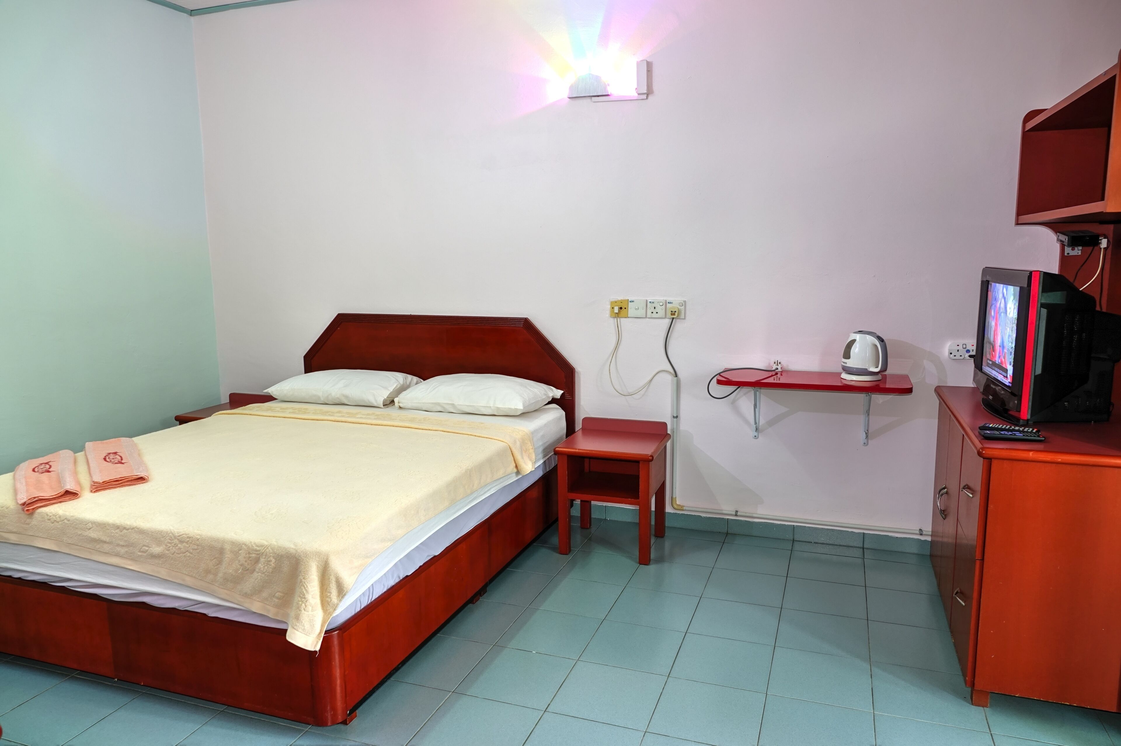 Une chambre d’hôtel avec un lit en bois, une petite table de chevet, une commode rouge, un téléviseur et une étagère fixée au mur.