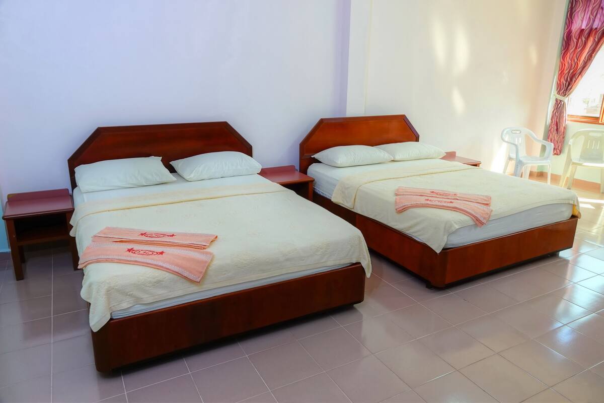Two wooden beds with white linens and pink towels, a small wooden nightstand, and a plastic chair near a window with curtains.