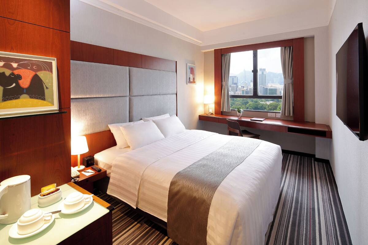 Habitación de hotel con una cama grande, un escritorio, una silla, un ventanal con vista a la ciudad y un televisor montado.
