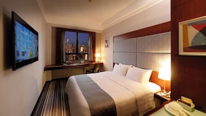 A hotel room with a bed, a desk, a chair, a TV, a window with a city view, and a nightstand with a lamp. - B P International (Kowloon)