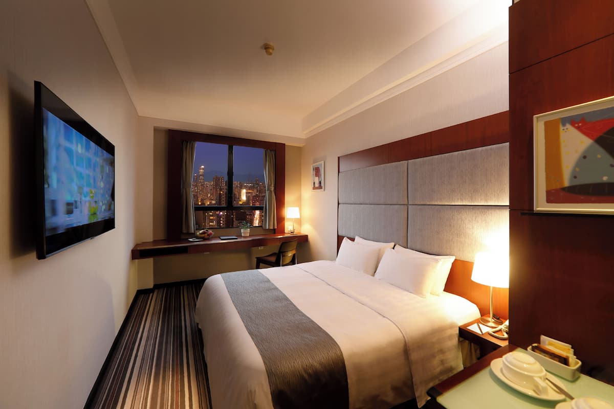 Una habitación de hotel con cama, escritorio, silla, televisión, ventana con vista a la ciudad y mesita de noche con lámpara.