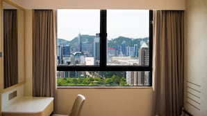 A hotel room with a large window offering a cityscape view, a white bedside table, and a chair. - B P International (Kowloon)