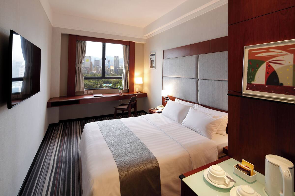 Habitación de hotel con una cama grande, un escritorio con silla, una ventana con vista a la ciudad y un cuadro en la pared.