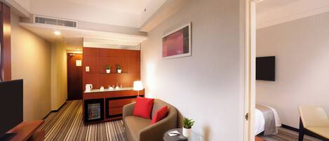 A hotel room with a sofa, a small table with a plant, a TV, a desk with a computer, and a bed.