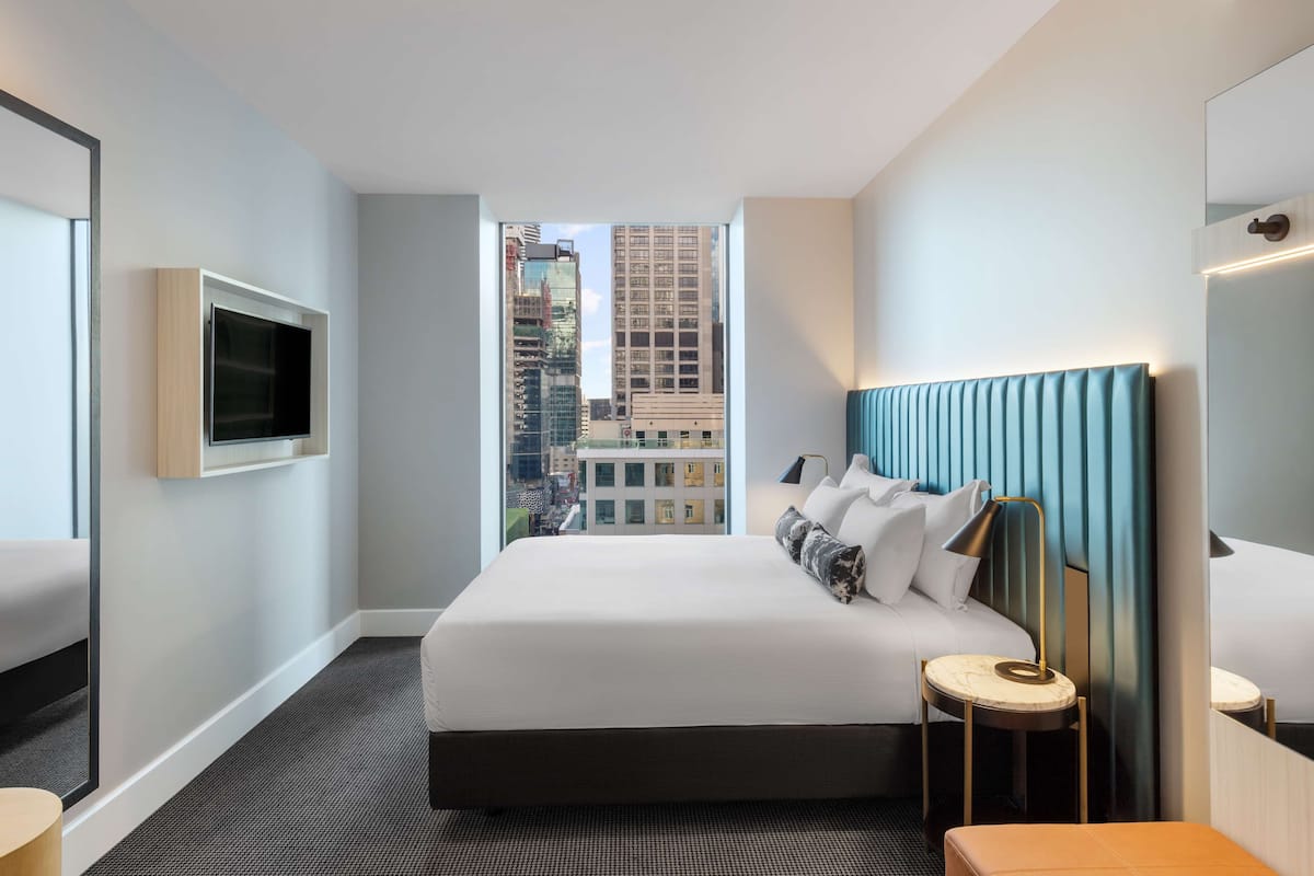 A hotel room with a large bed, a TV mounted on the wall, a bedside table, and a view of the cityscape through the window.