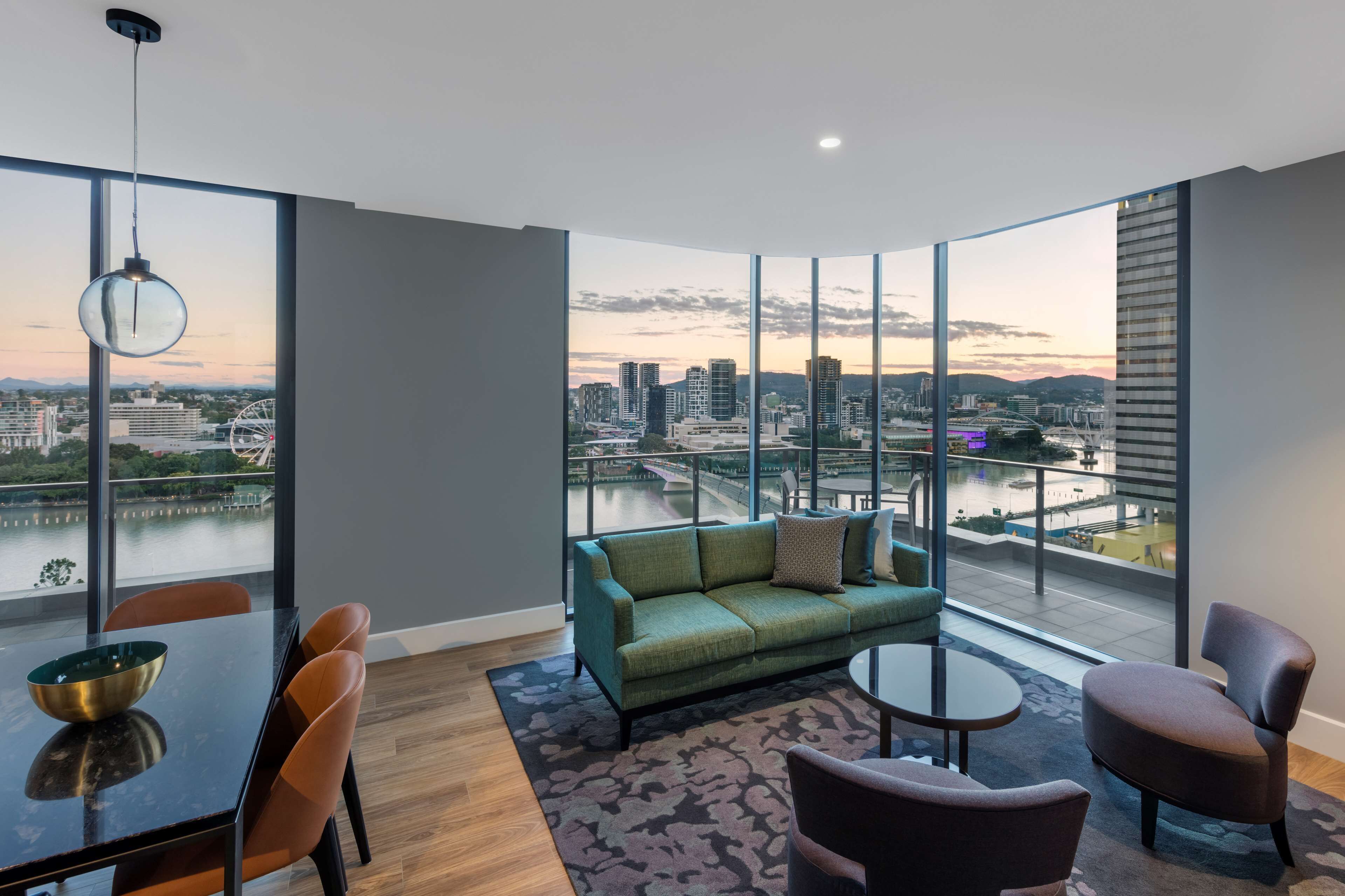 A modern living room with a green sofa, a dining table with chairs, and a view of a cityscape and river.
