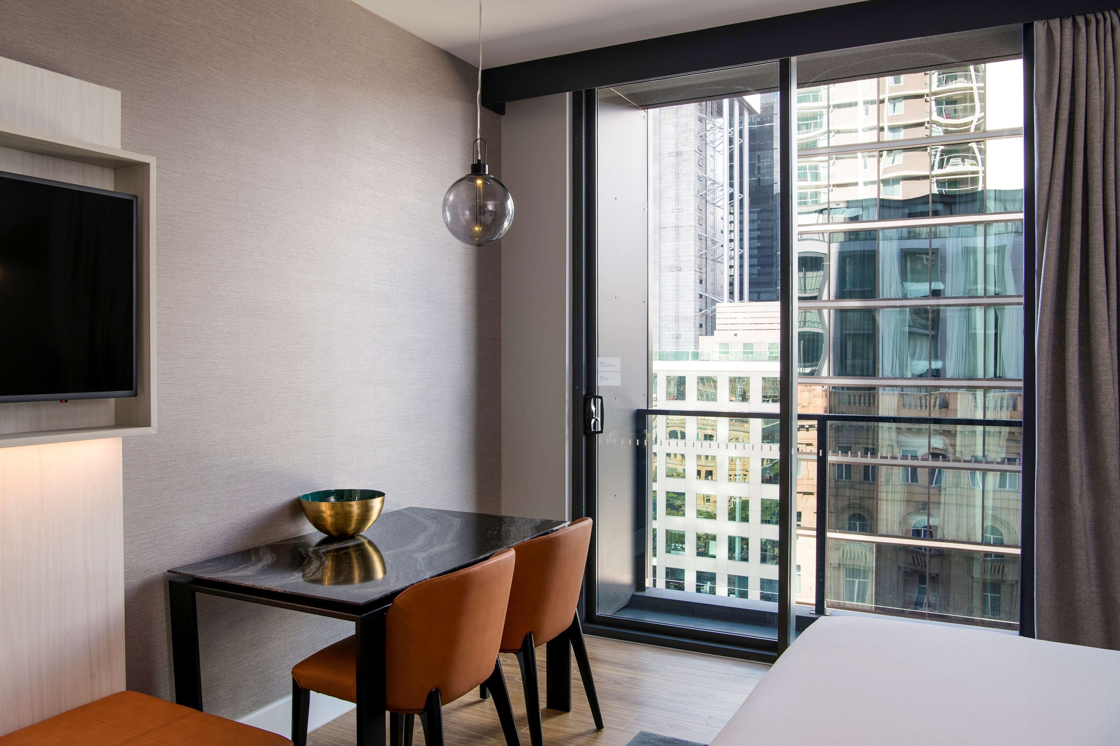 Une chambre d’hôtel moderne dotée d’une grande fenêtre, d’un téléviseur à écran plat, d’une table à manger avec des chaises et d’un petit vase décoratif.