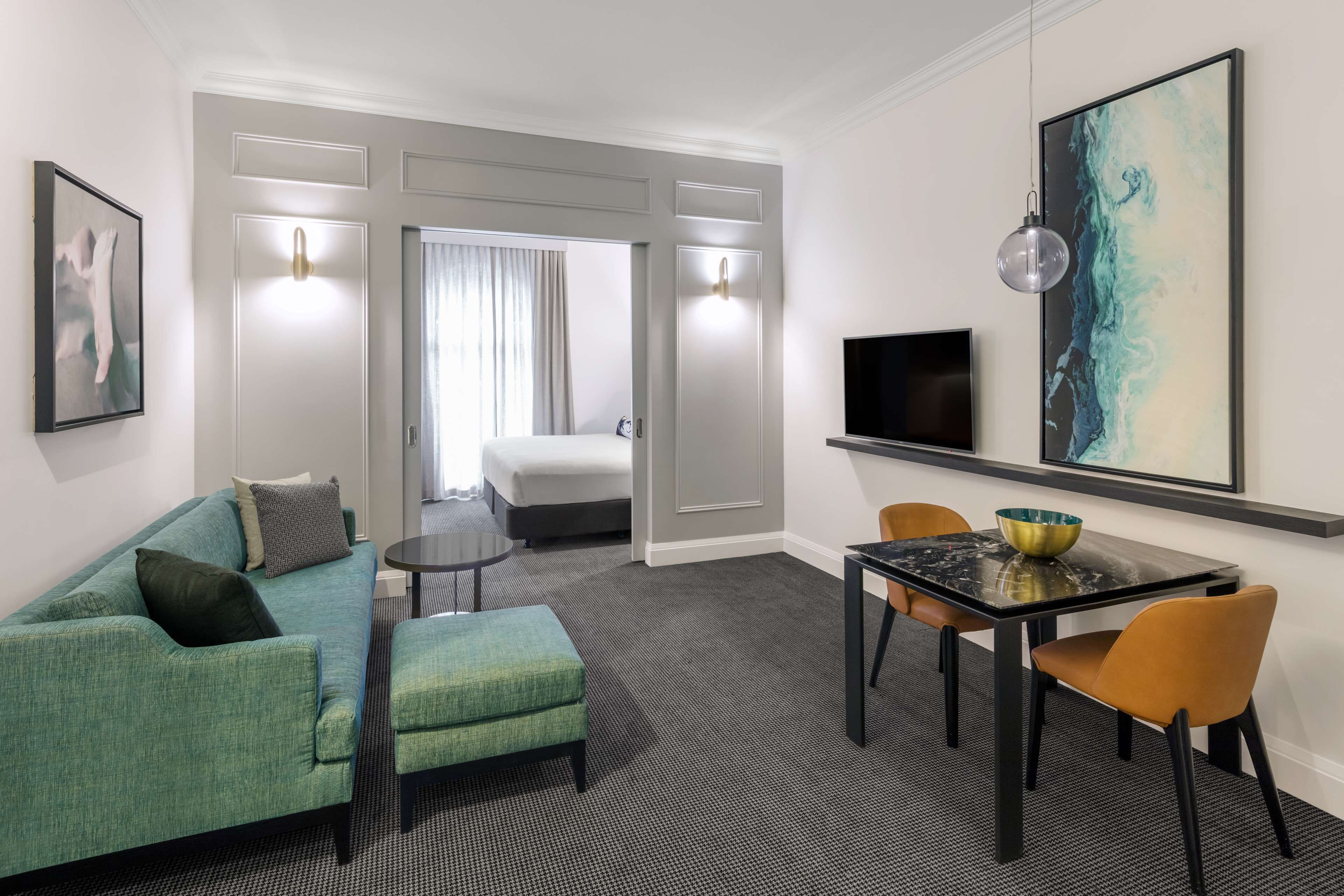 Habitación de hotel con un sofá verde, una mesita con un frutero, un televisor y un cuadro en la pared.