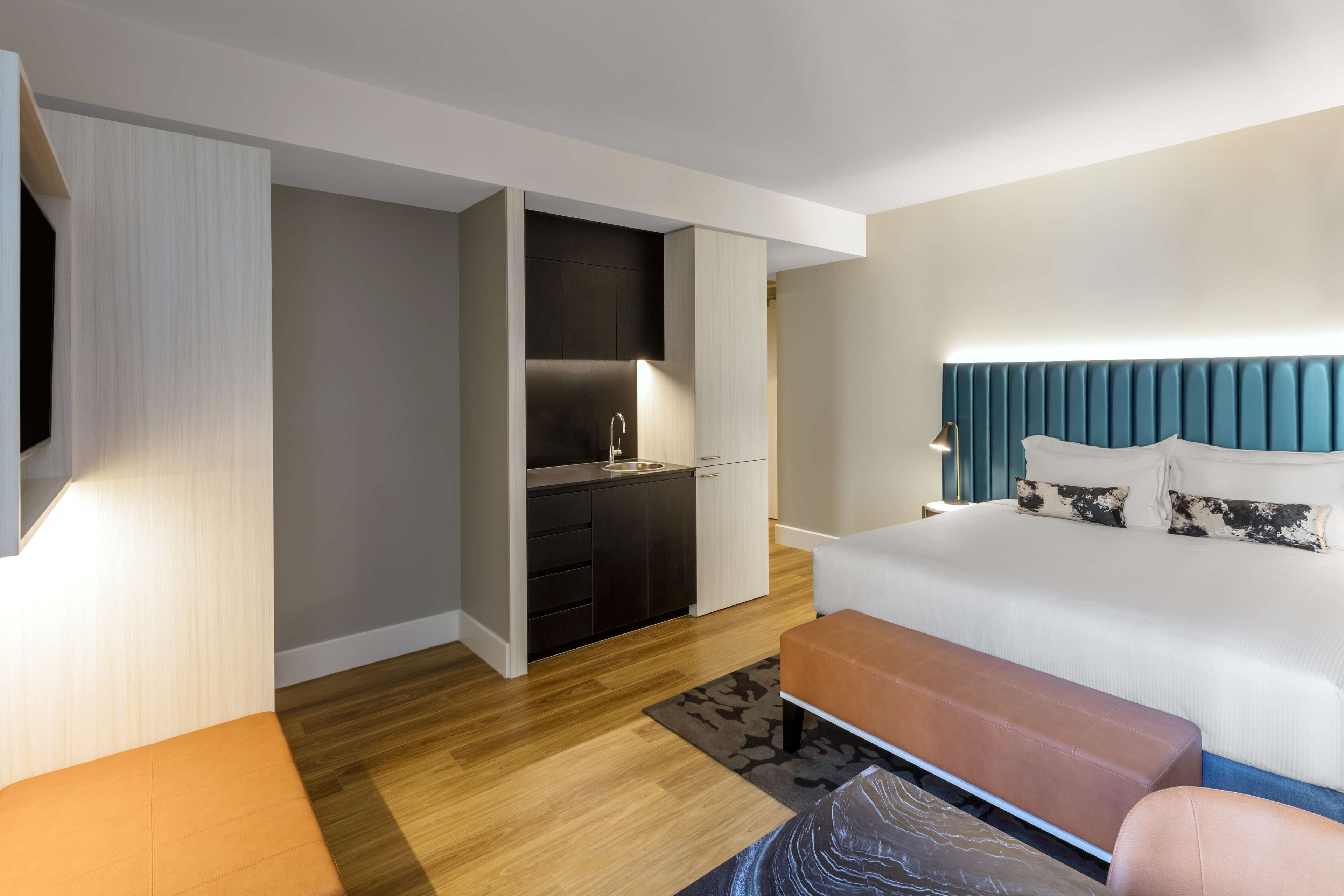 A modern hotel room with a bed, a small kitchenette, and a seating area.