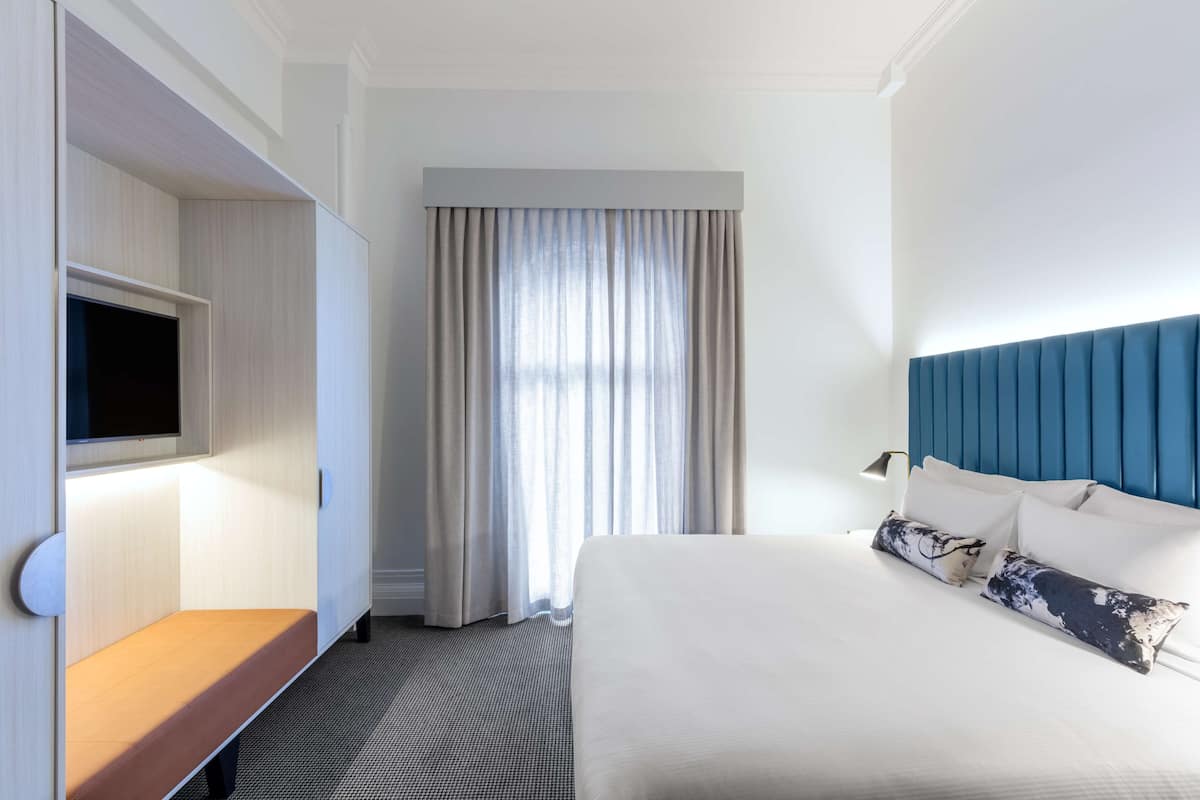 A hotel room with a bed, a TV mounted on a cabinet, and a large window with curtains.