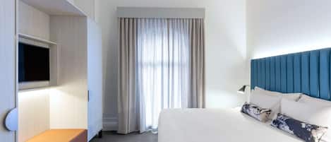Una habitación de hotel con una cama, un televisor en un mueble y un ventanal con cortinas.