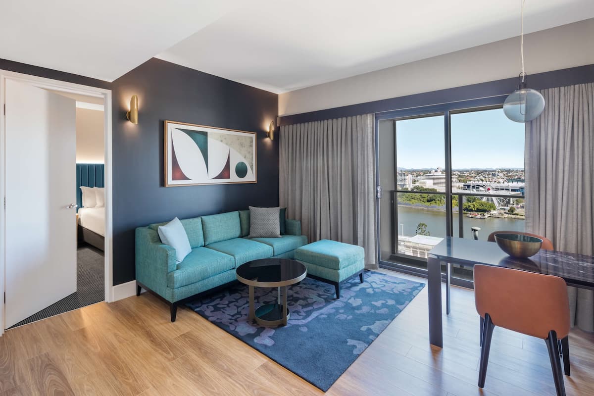 A modern living room with a teal sofa, a round coffee table, and a dining area with a table and chairs. A large window offers a view of a cityscape.