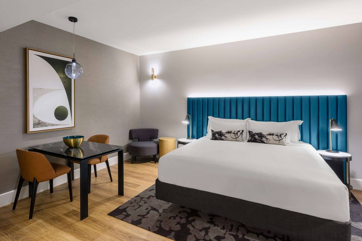 A modern hotel room with a large bed, a dining table, and a seating area.