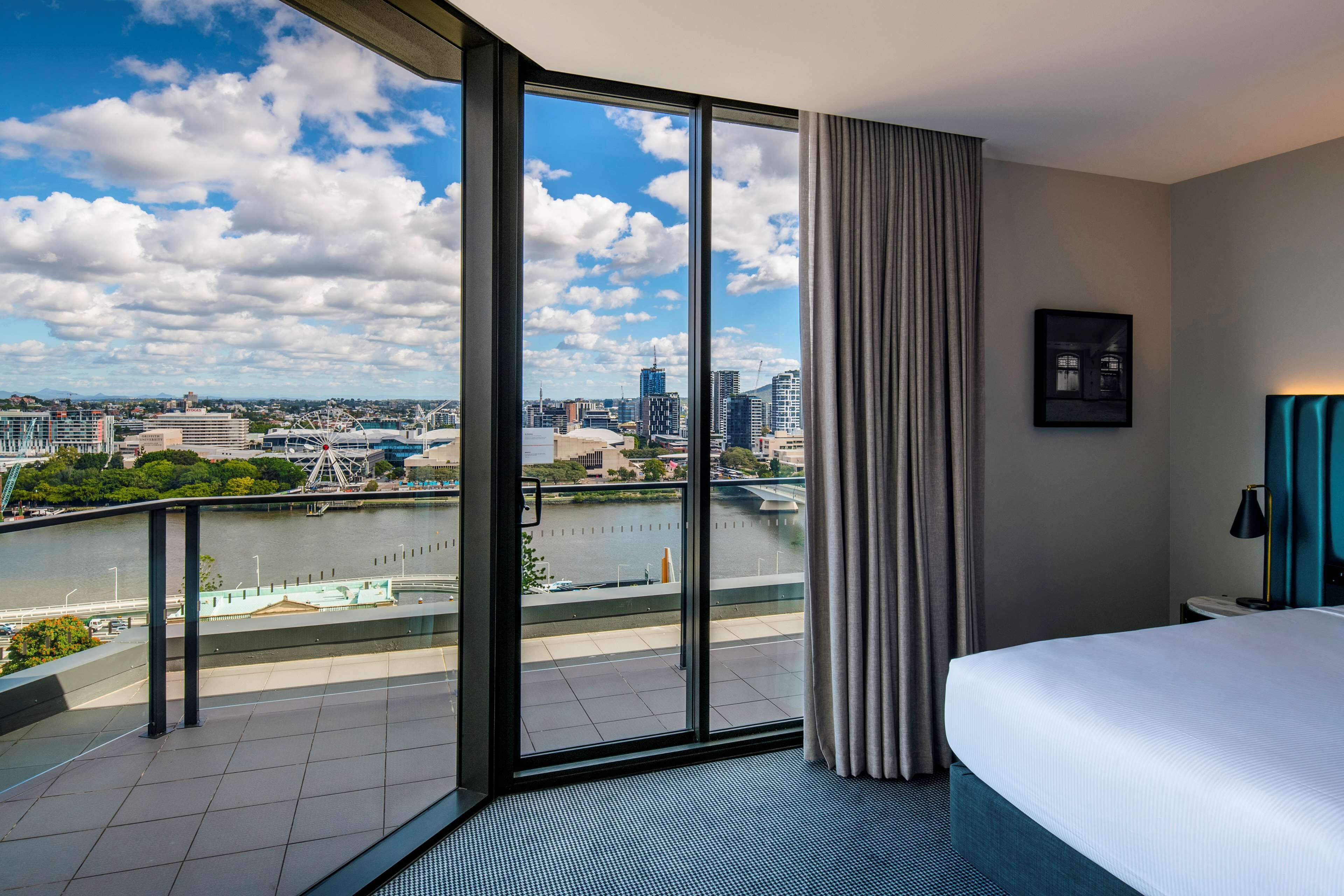 Een moderne hotelkamer met een groot raam met uitzicht over de stad, een bed, een nachtkastje en een rivier met een brug.