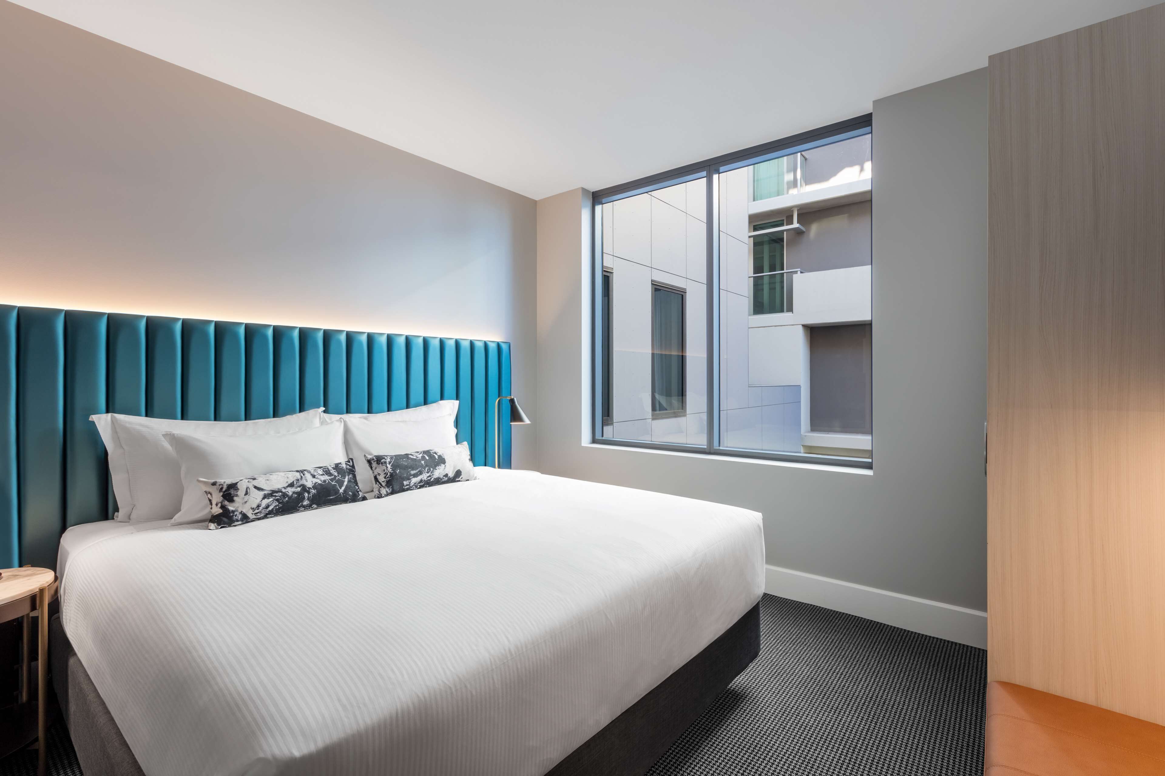 Une chambre d’hôtel moderne avec un grand lit, une tête de lit bleue, une fenêtre donnant sur le bâtiment et un plancher en bois.