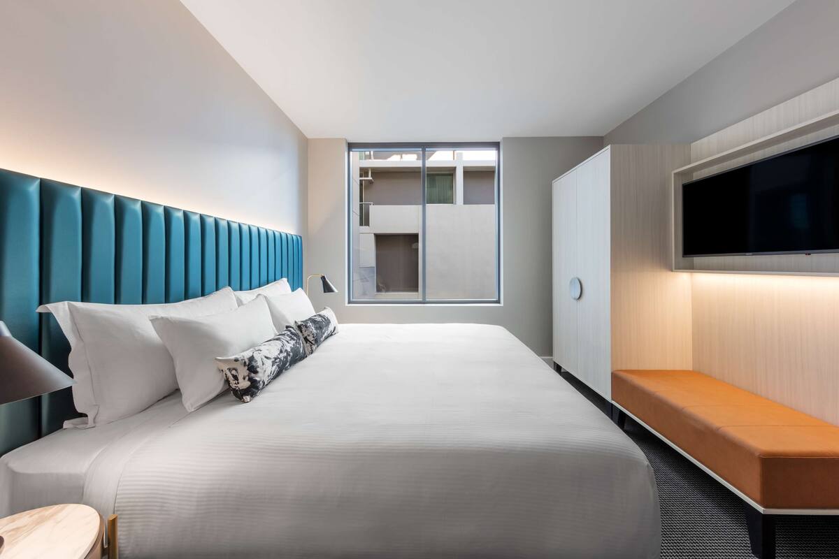 A modern hotel room with a large bed, a TV mounted on the wall, and a view of the building's interior through a glass door.