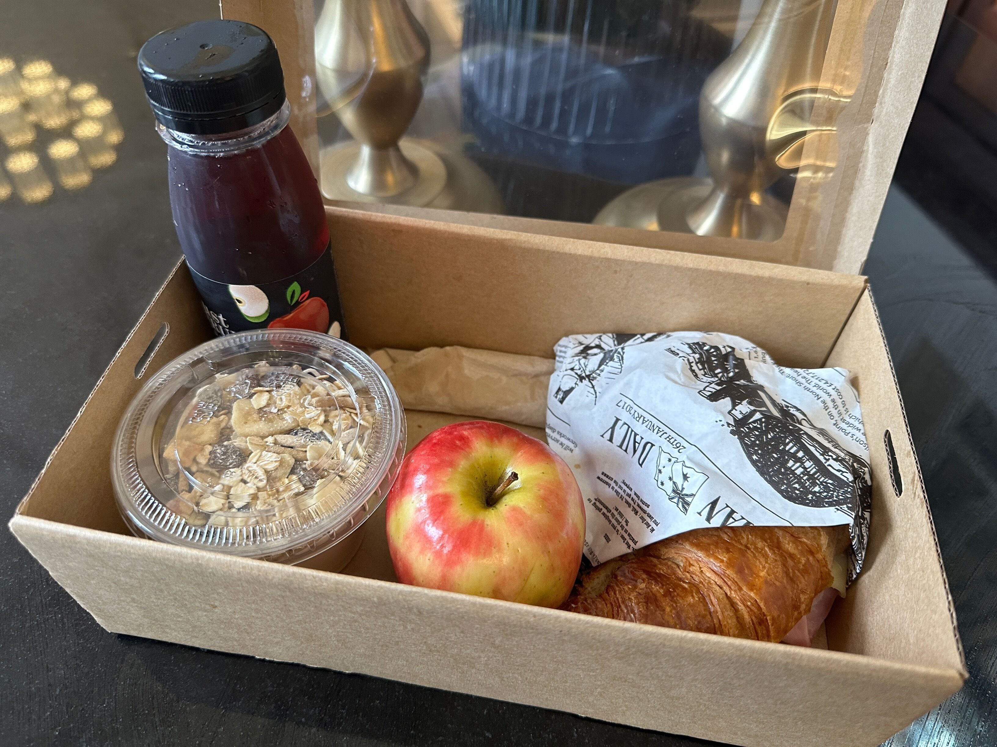 Una scatola di cartone con un succo di frutta, un contenitore di plastica con cereali, una mela e un croissant.