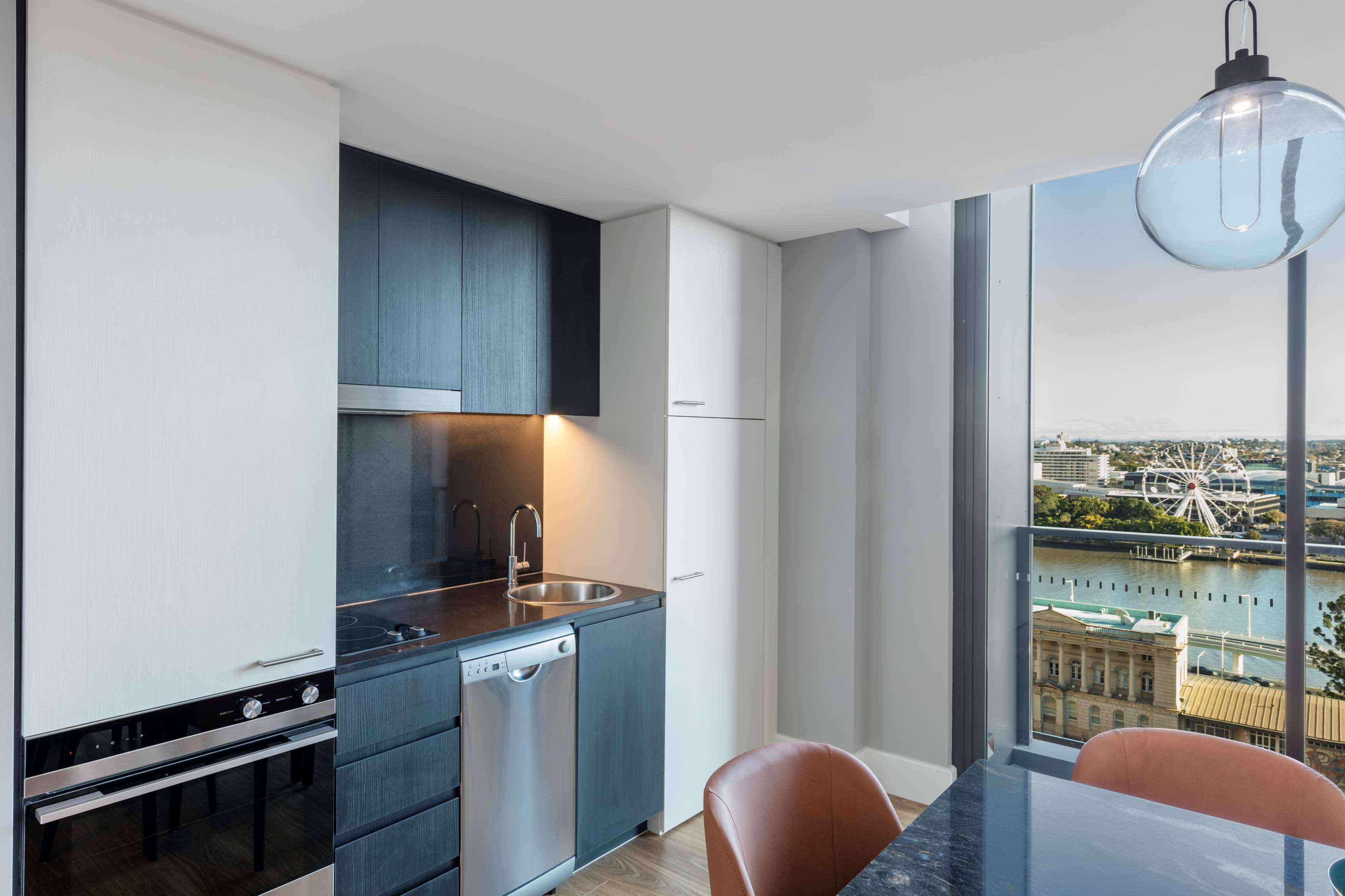 A modern kitchen with a stainless steel sink, a dishwasher, and a built-in oven. There is a dining area with a table and chairs, and a large window offering a view of a cityscape.
