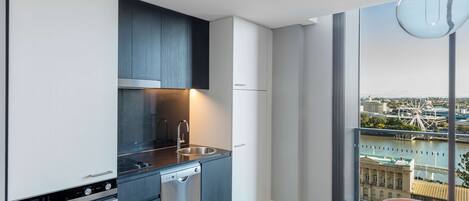 Una cocina moderna con un lavaplatos de acero inoxidable, lavavajillas y horno empotrado. Hay una zona de comedor con una mesa y sillas, y un amplio ventanal con vistas a la ciudad.