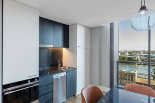 A modern kitchen with a stainless steel sink, a dishwasher, and a built-in oven. There is a dining area with a table and chairs, and a large window offering a view of a cityscape.