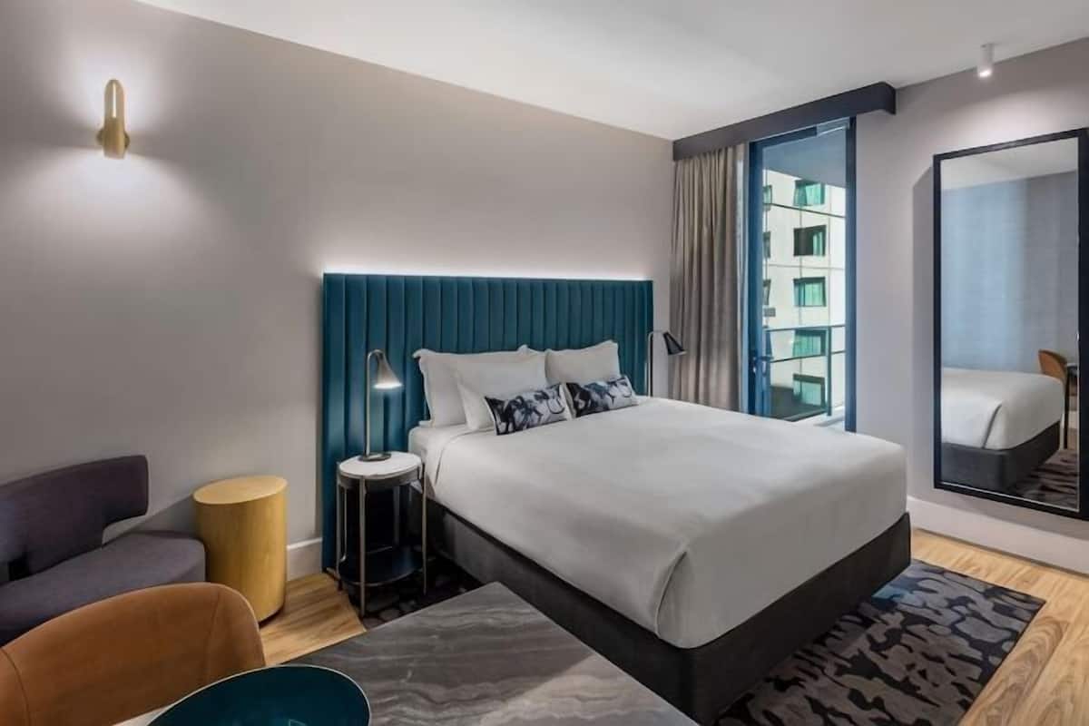A modern hotel room with a large bed, a desk, a chair, a round side table, and a large mirror.