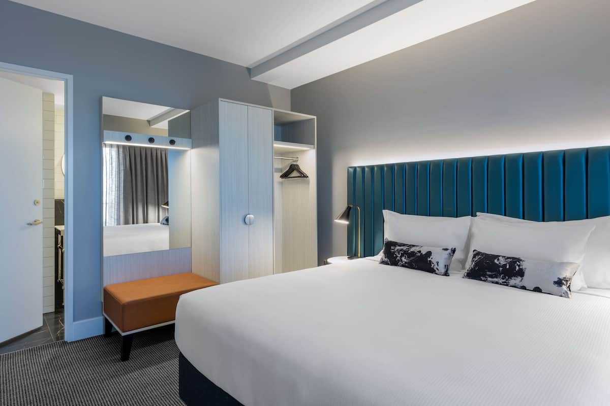 A modern hotel room with a large bed, a bench, a walk-in closet, and a bathroom visible through an open door.