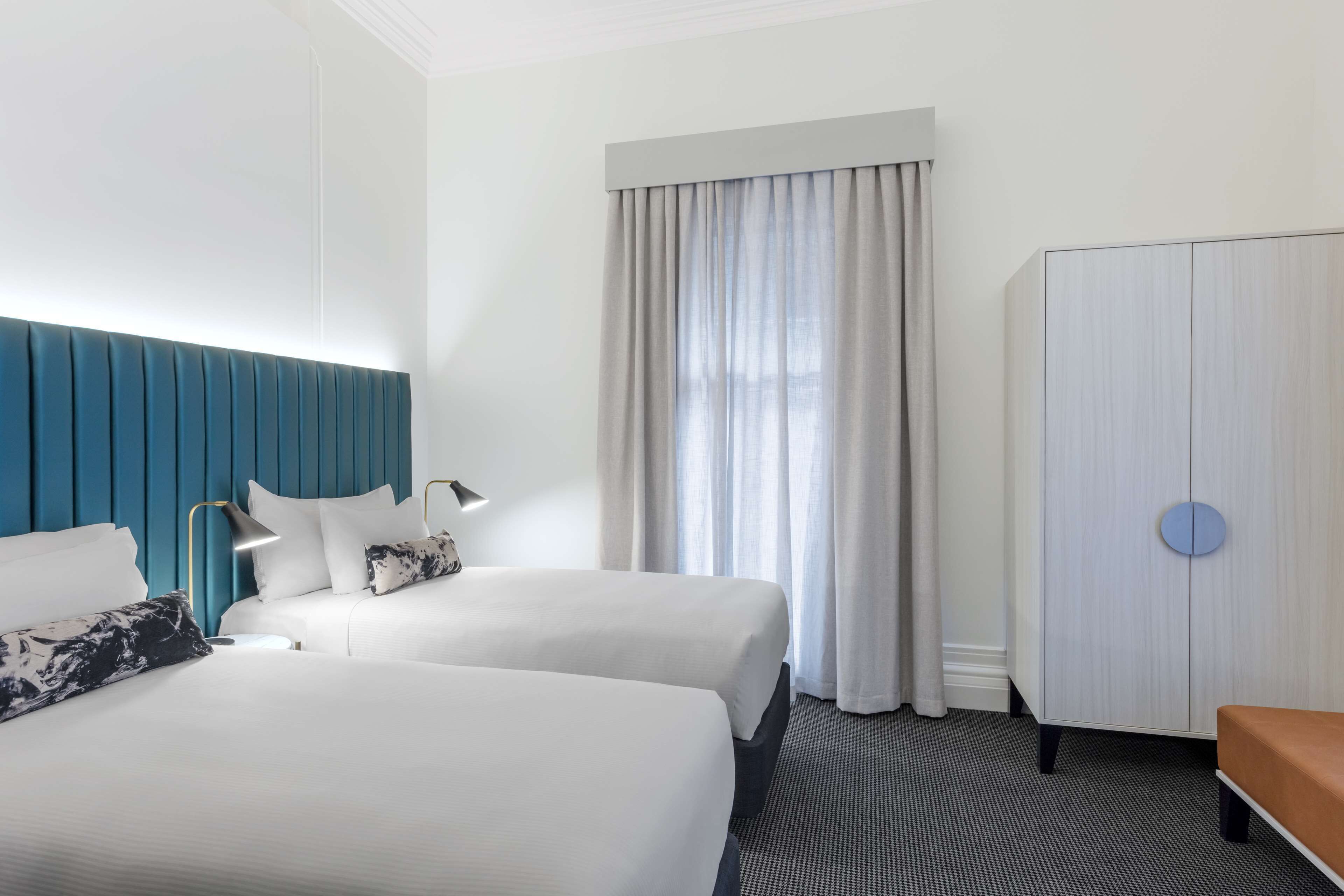 A hotel room with two beds, a blue headboard, a wardrobe, and a window with curtains.