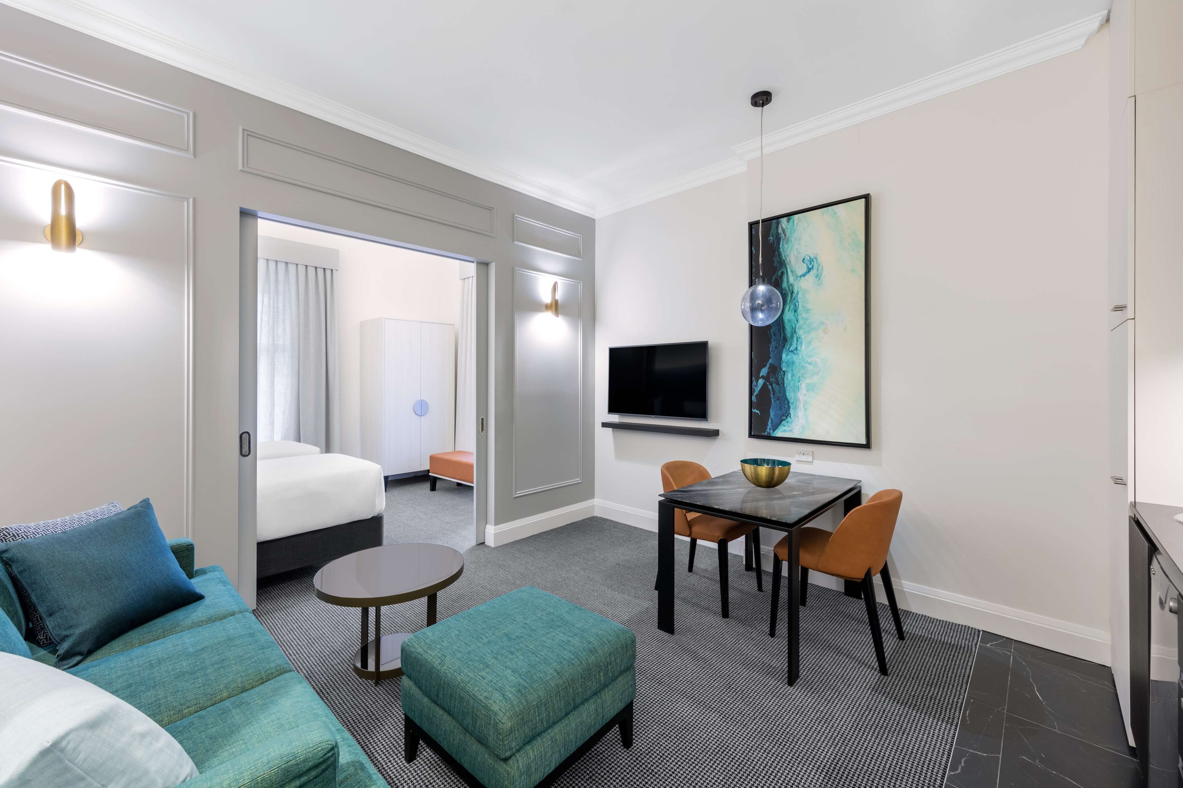 Een hotelkamer met een bank, voetenbankje, eettafel, stoelen, een televisie en kunstwerken aan de muur.