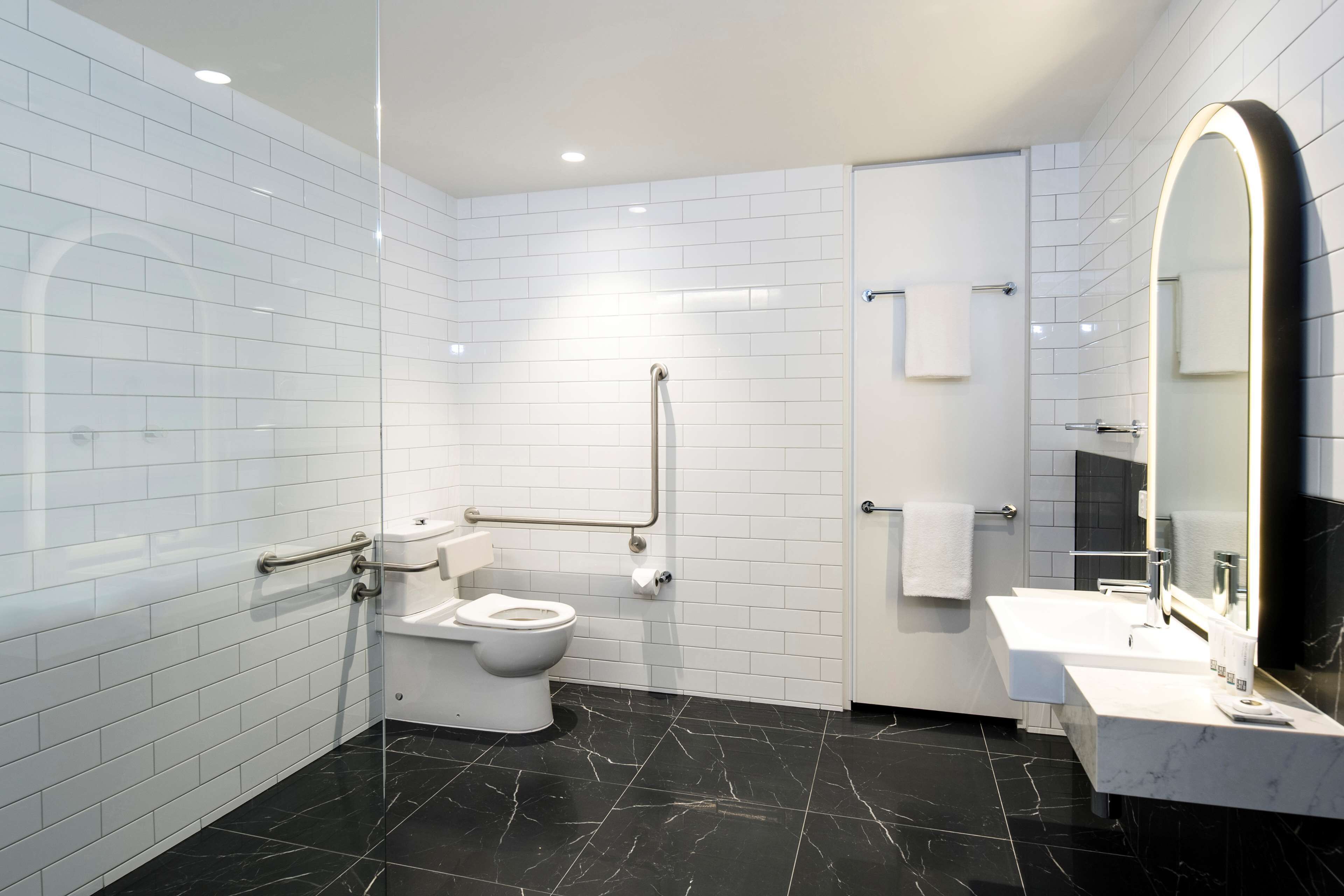 A modern bathroom with white tiled walls, a black marble floor, a white toilet, a sink with a mirror, and a handrail.