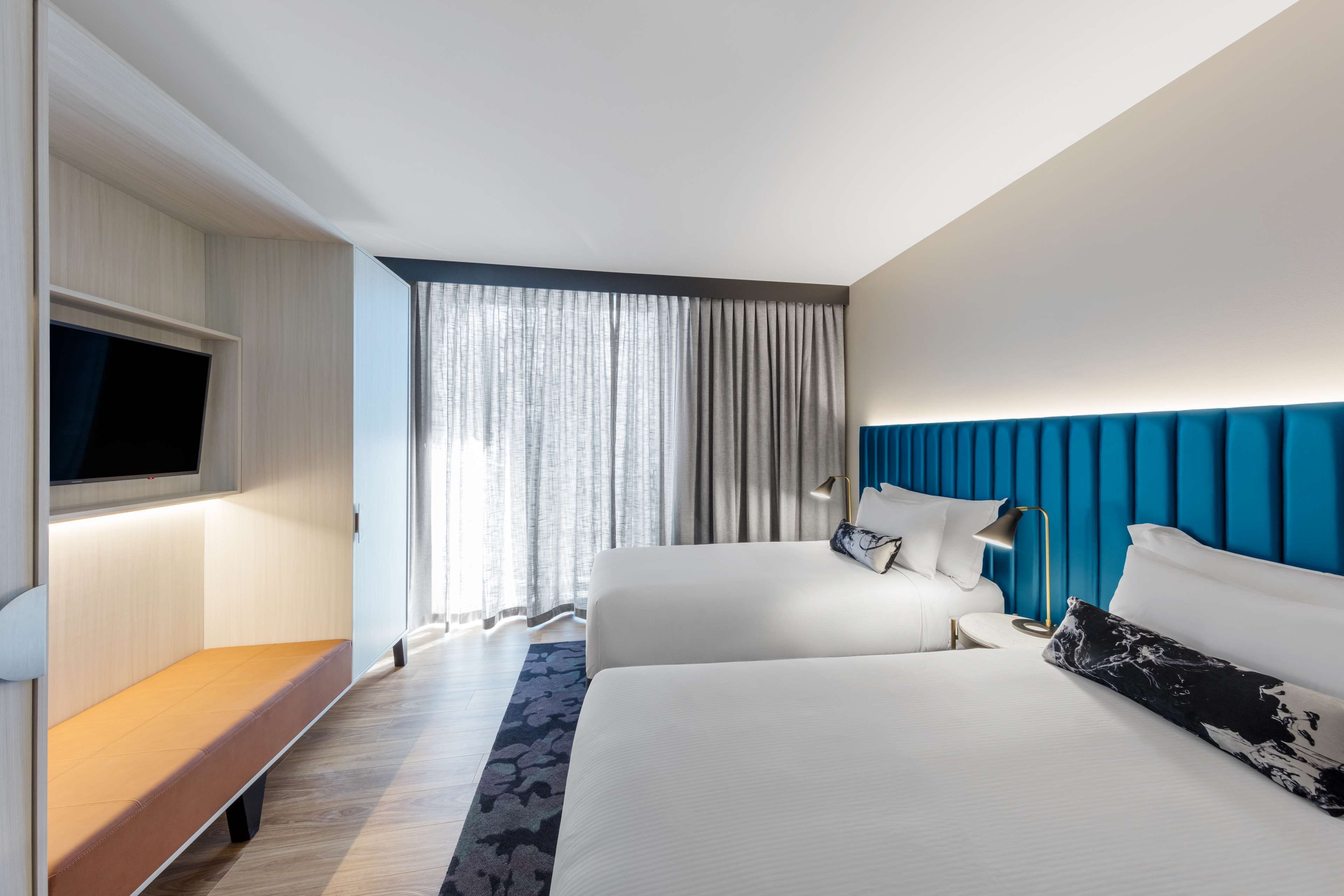 Una habitación de hotel moderna con una cama grande, una cama más pequeña, un televisor de pantalla plana, un banco y un amplio ventanal con cortinas.
