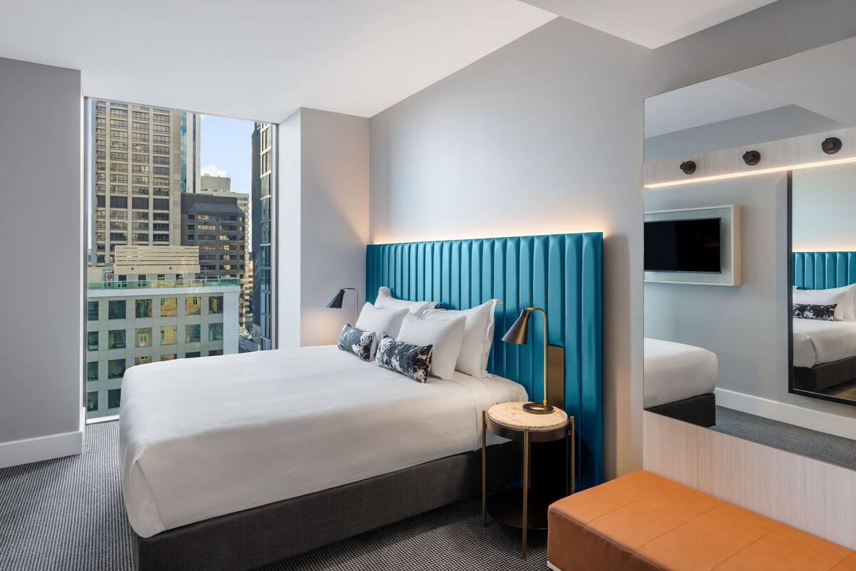 A modern hotel room with a large bed, a bedside table, a mirror, and a city view.