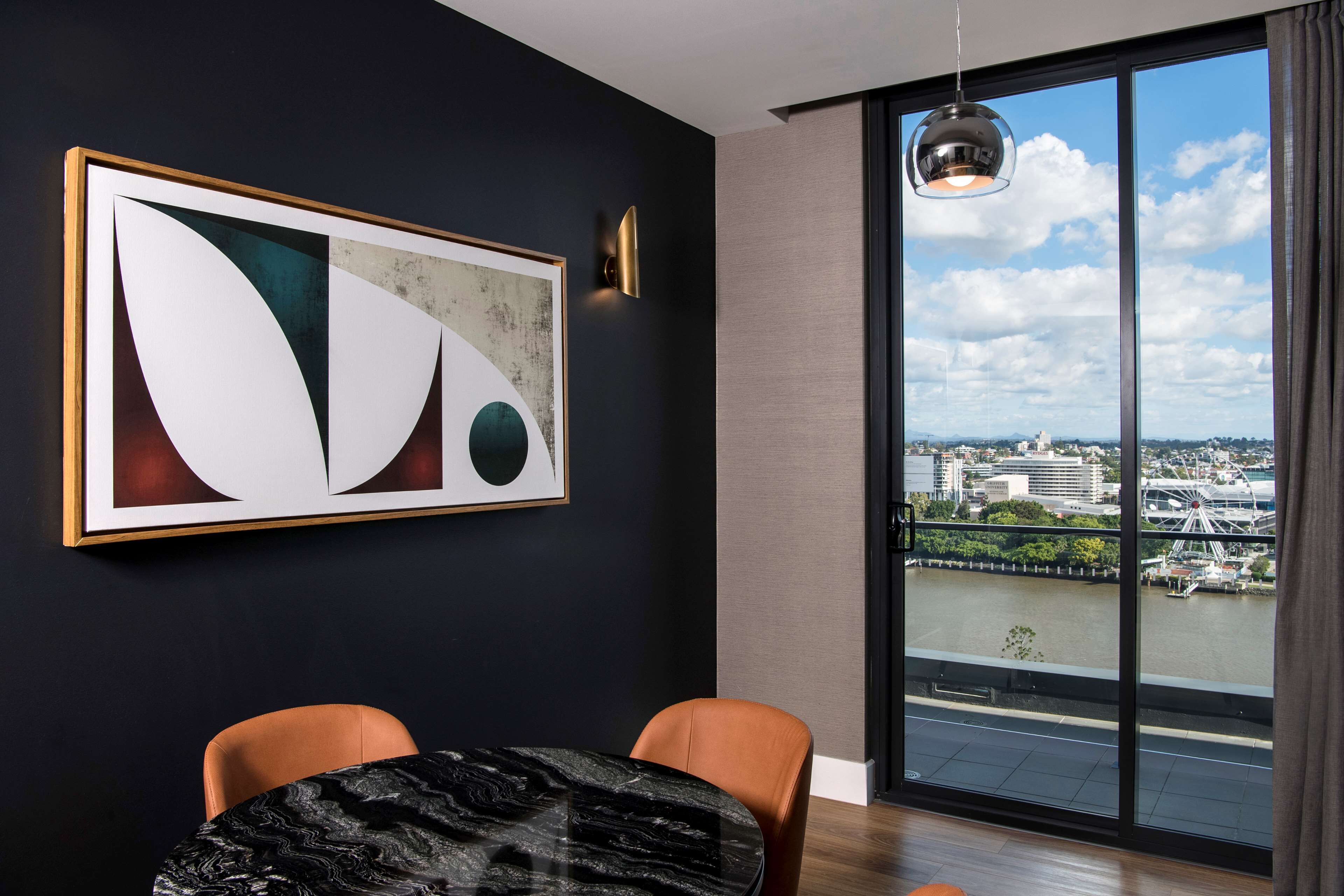 Un comedor moderno con una mesa de mármol, dos sillas, un cuadro abstracto de gran tamaño y vistas a un paisaje urbano a través de amplias ventanas.
