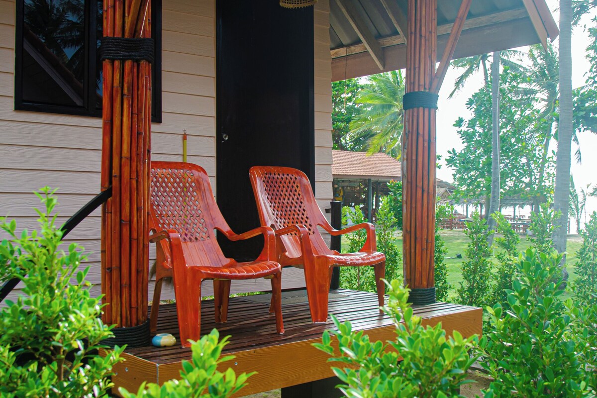 A wooden porch with two red plastic chairs and a bench, surrounded by lush greenery.