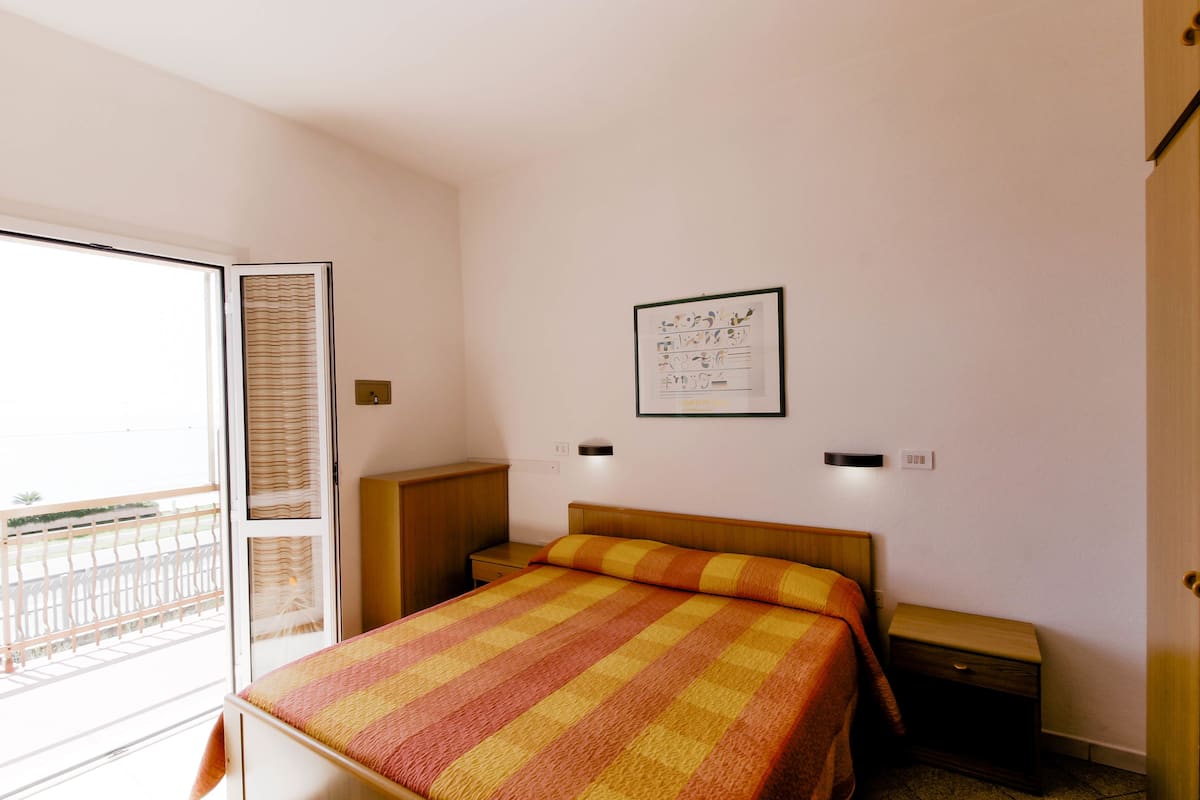 A bedroom with a bed, a nightstand, a wall-mounted light, a window with blinds, and a framed picture on the wall.