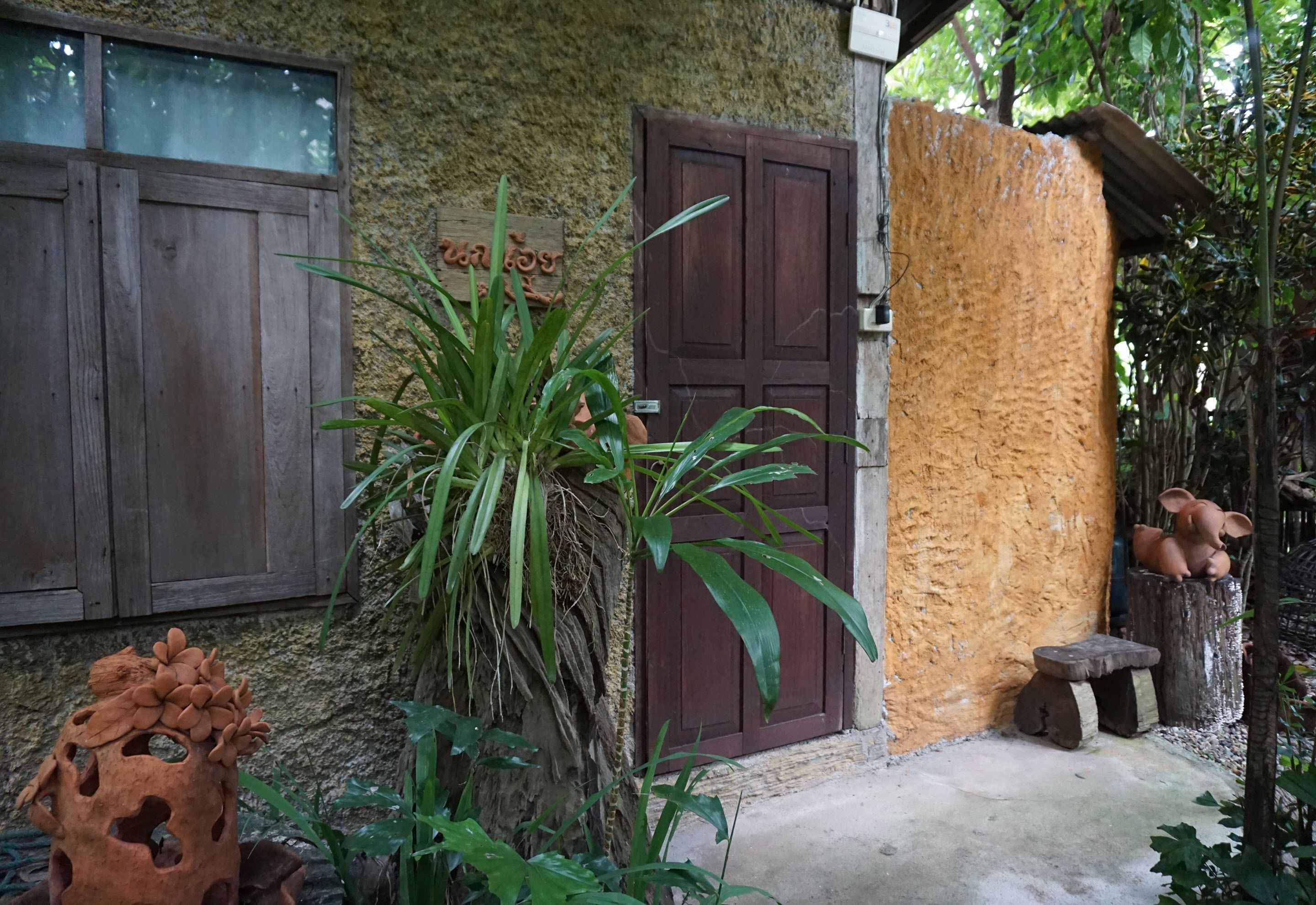 Un edificio rústico con puerta de madera, una planta grande en maceta y un banco de madera.