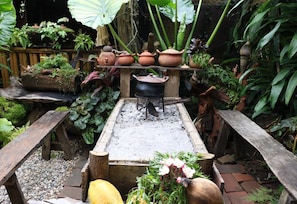 Un entorno rústico al aire libre con un banco de madera, un pequeño hogar para fuego y diversas plantas en macetas.