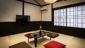Une pièce de style japonais traditionnel, avec un sol recouvert de tatamis, une petite table sur laquelle sont posés un vase et une théière, un téléviseur fixé au mur et un paravent en papier de riz (shoji).