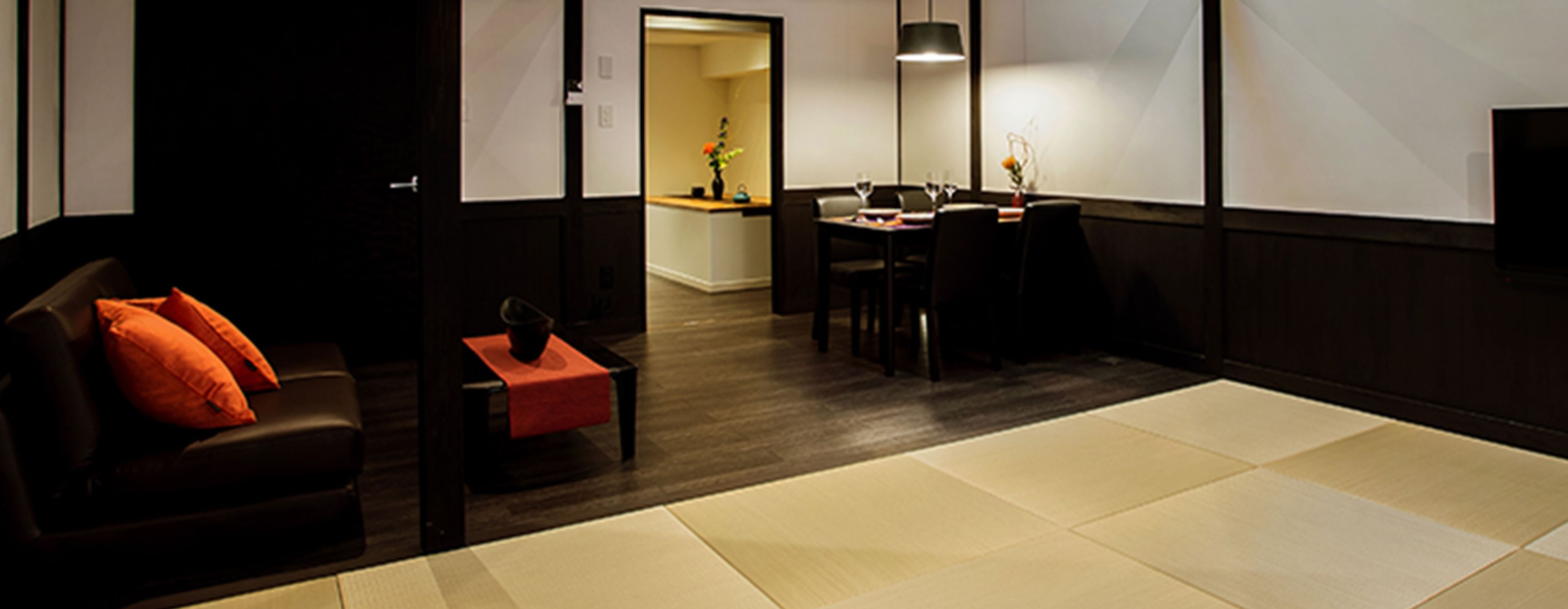 A modern hotel room with a dark wood floor, a sofa with orange cushions, a small table with a vase, and a dining area with a table and chairs.