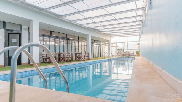 Una piscina interior con techos de vidrio transparente, sillas junto a la piscina y suelo de baldosas.