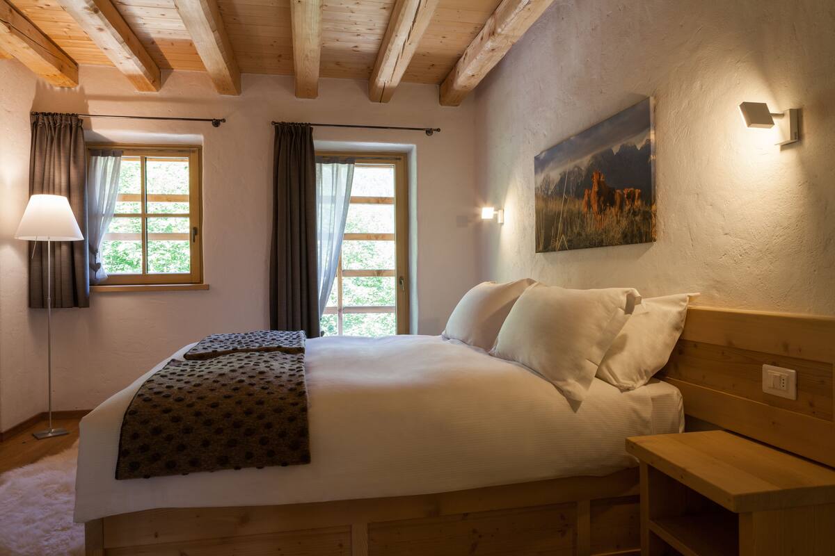 A bedroom with a large bed, wooden headboard, two windows with curtains, a painting on the wall, and a bedside table.