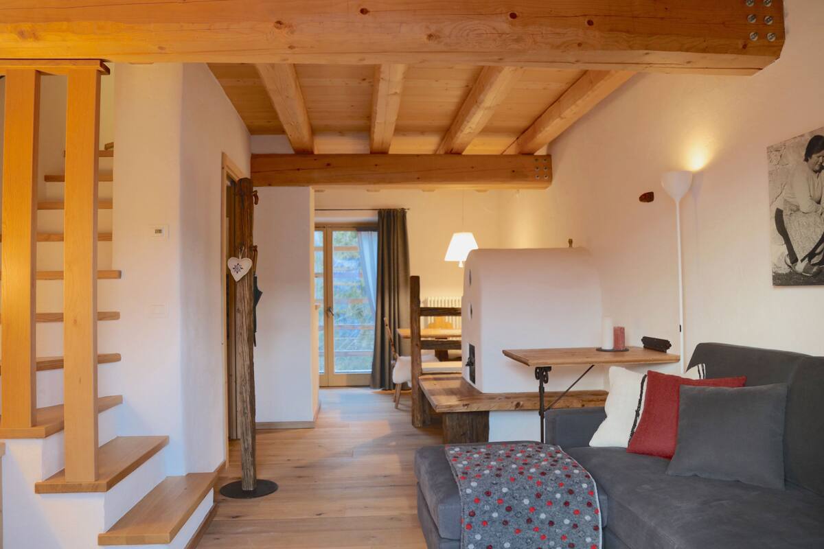 A cozy room with a wooden staircase, a sofa with patterned throw, a table with a lamp, and a view through a window.
