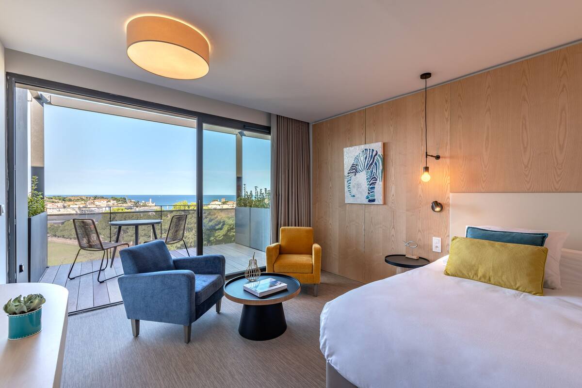 Een moderne hotelkamer met een groot bed, een bank, een stoel, een tafeltje en uitzicht op de stad door grote ramen.
