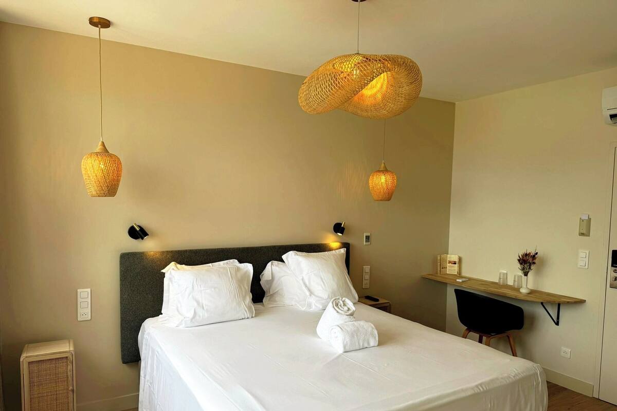 Een netjes opgemaakt bed met witte lakens, twee bedlampjes en een klein nachtkastje met een afstandsbediening.