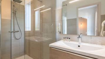 Un baño moderno con ducha envuelta en vidrio, un espejo grande y un lavabo blanco con artículos de aseo.