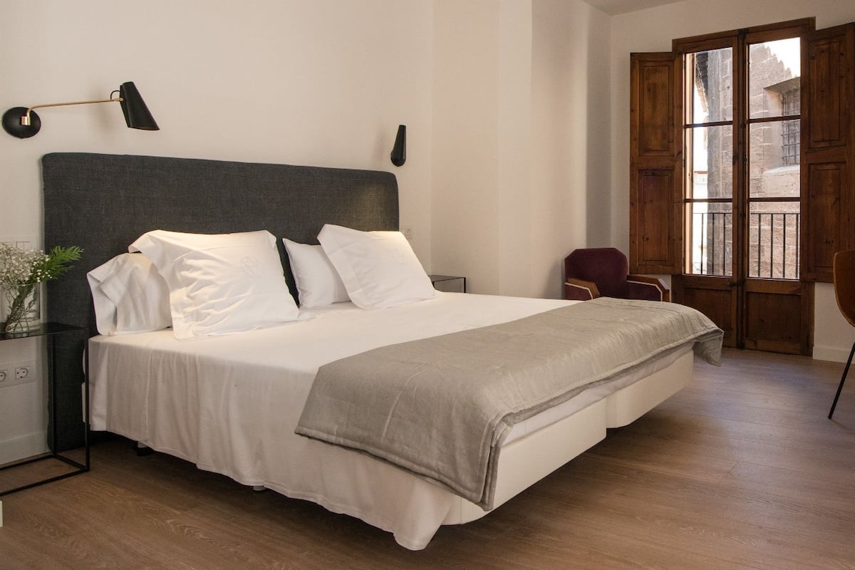 A neatly made bed with white linens, a dark headboard, and a grey throw blanket. There is a desk with a chair and a lamp to the right.