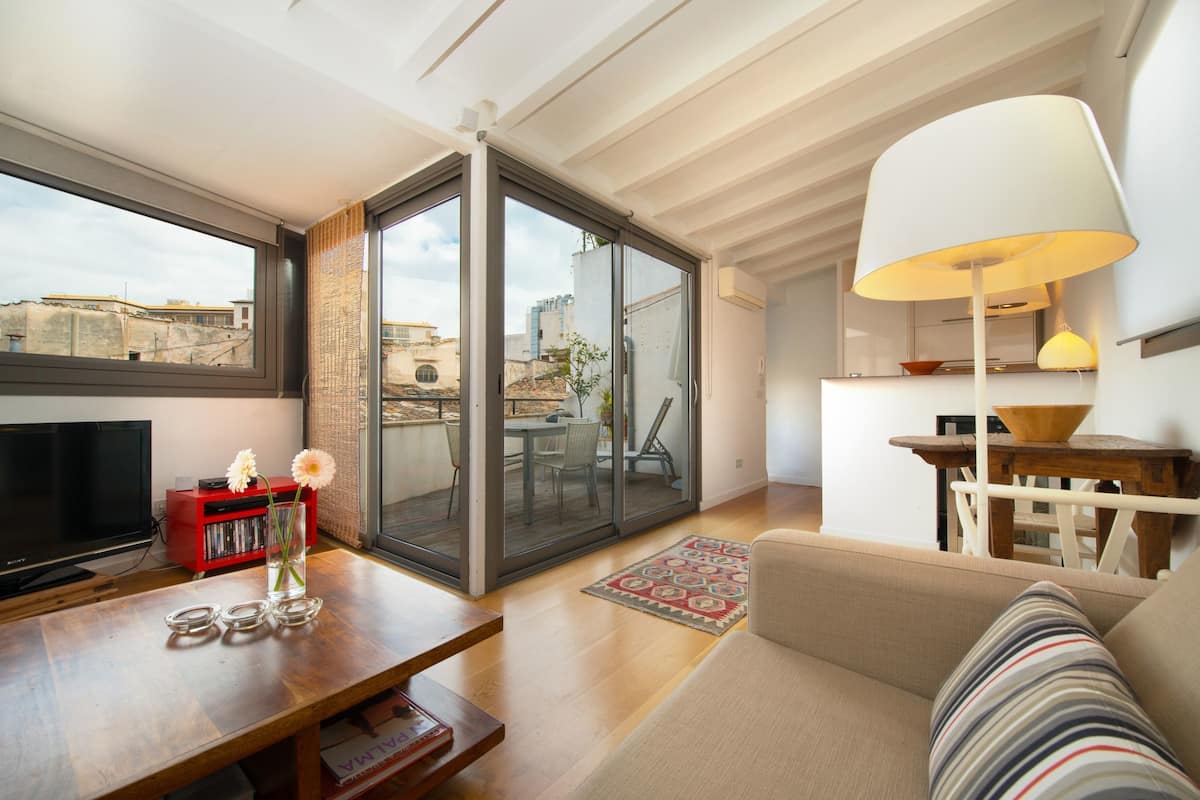 A modern living room with a sofa, a TV, a bookshelf, and a large window offering a city view.
